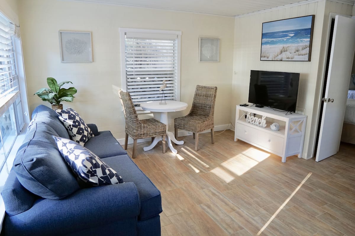 Un salon chaleureux avec un canapé bleu, des fauteuils en osier, une petite table, un téléviseur et un tableau sur le thème de la plage.