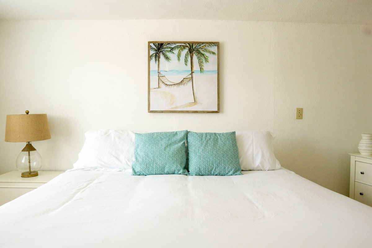 Un lit bien fait, avec du linge de lit blanc et deux coussins turquoise, une petite table de chevet avec une lampe, et une œuvre d’art encadrée représentant une scène de plage accrochée au mur.
