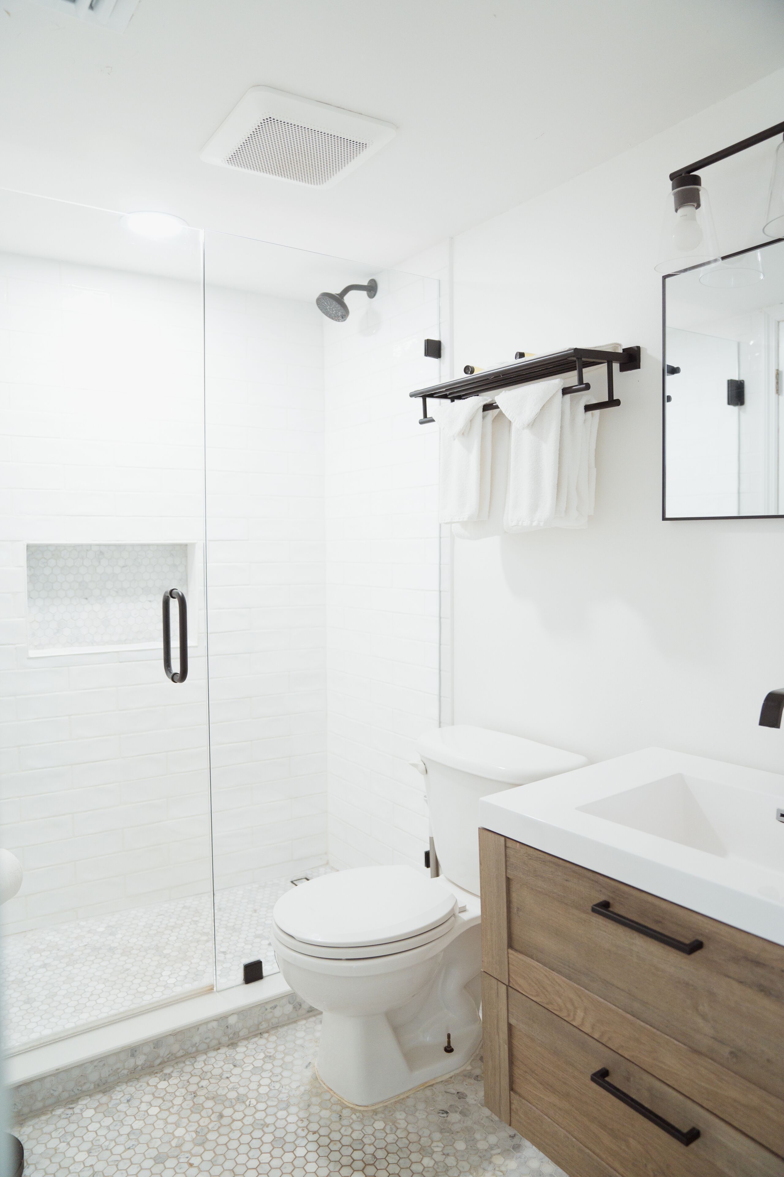 Un baño moderno con cabina de ducha de vidrio, inodoro blanco, tocador de madera con cajones y un espejo grande sobre el lavabo.