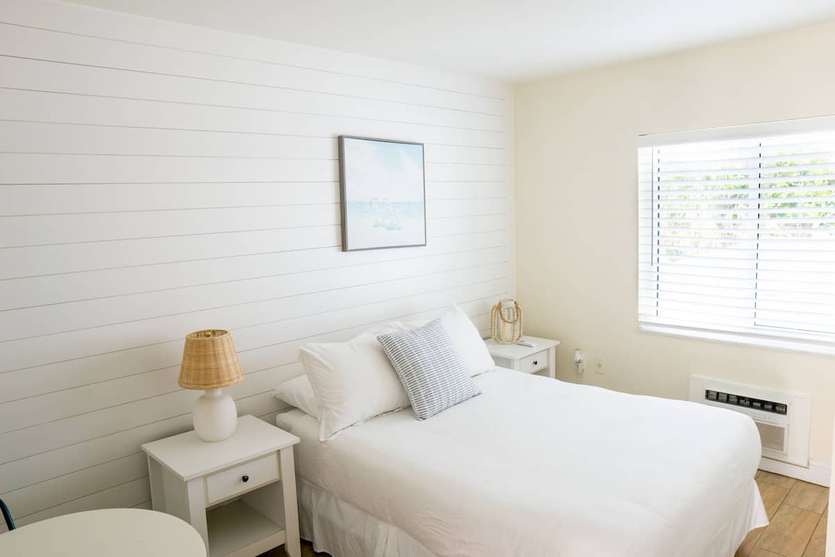 Une chambre à coucher avec un lit, une table de chevet avec une lampe, une commode, une fenêtre avec des stores et un tableau accroché au mur.