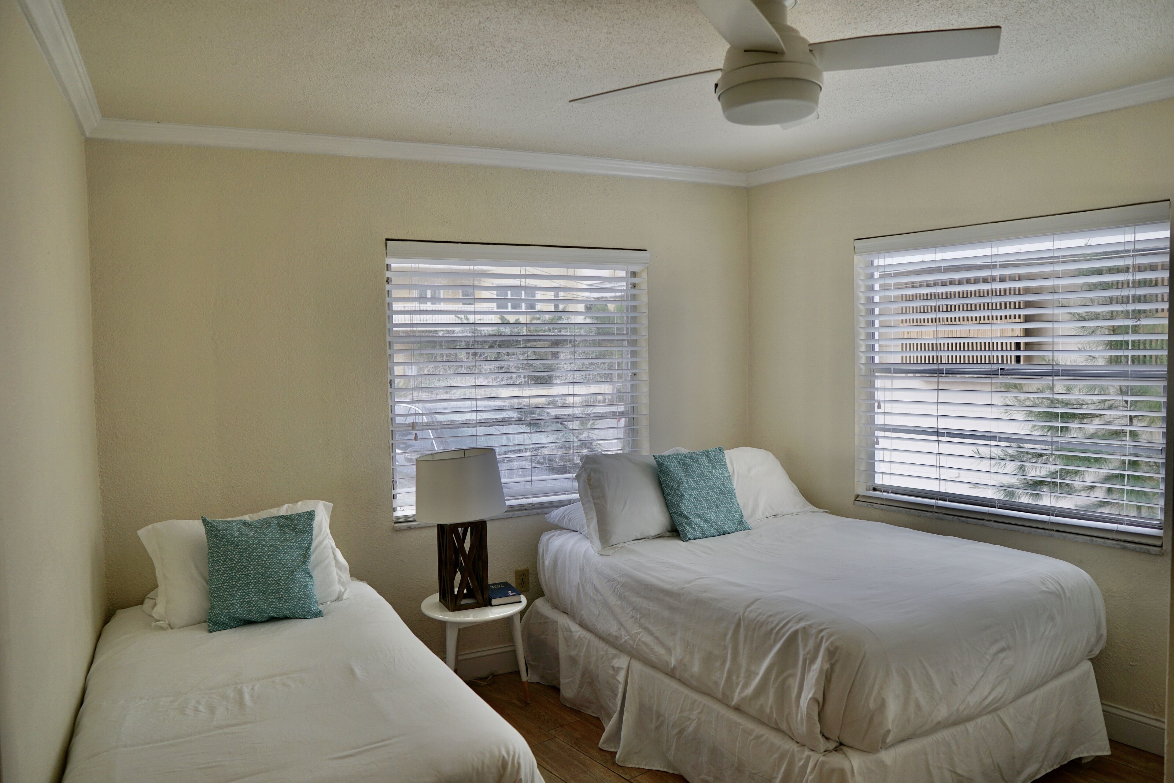 Quarto com duas camas, ventilador de teto e duas janelas com cortinas.