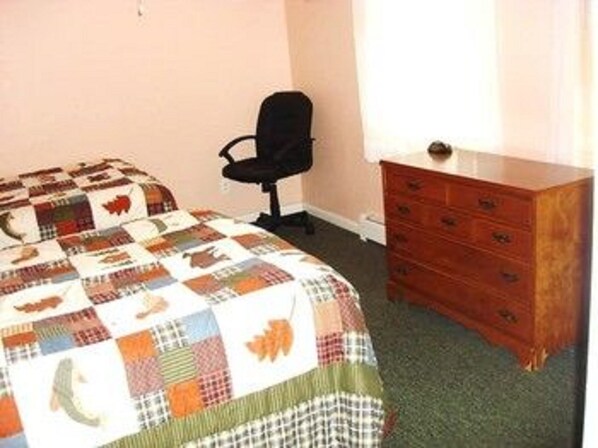 A bedroom with a bed covered in a patchwork quilt, a wooden dresser, and a black office chair. - Patio Motor Court (Twin Mountain)