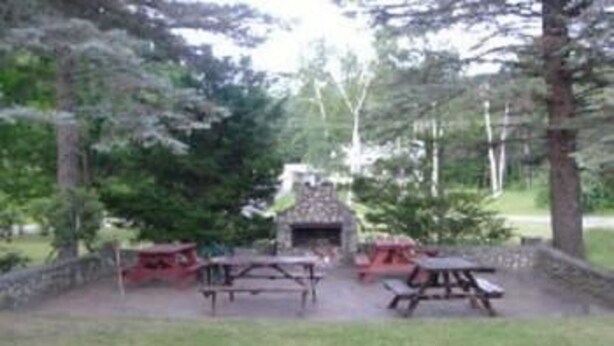 A rustic outdoor seating area with picnic tables and a stone fireplace surrounded by trees. - Patio Motor Court (Twin Mountain)