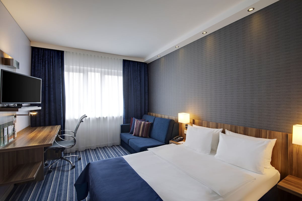 A hotel room with a bed, a desk, a chair, a TV, and a window with curtains.