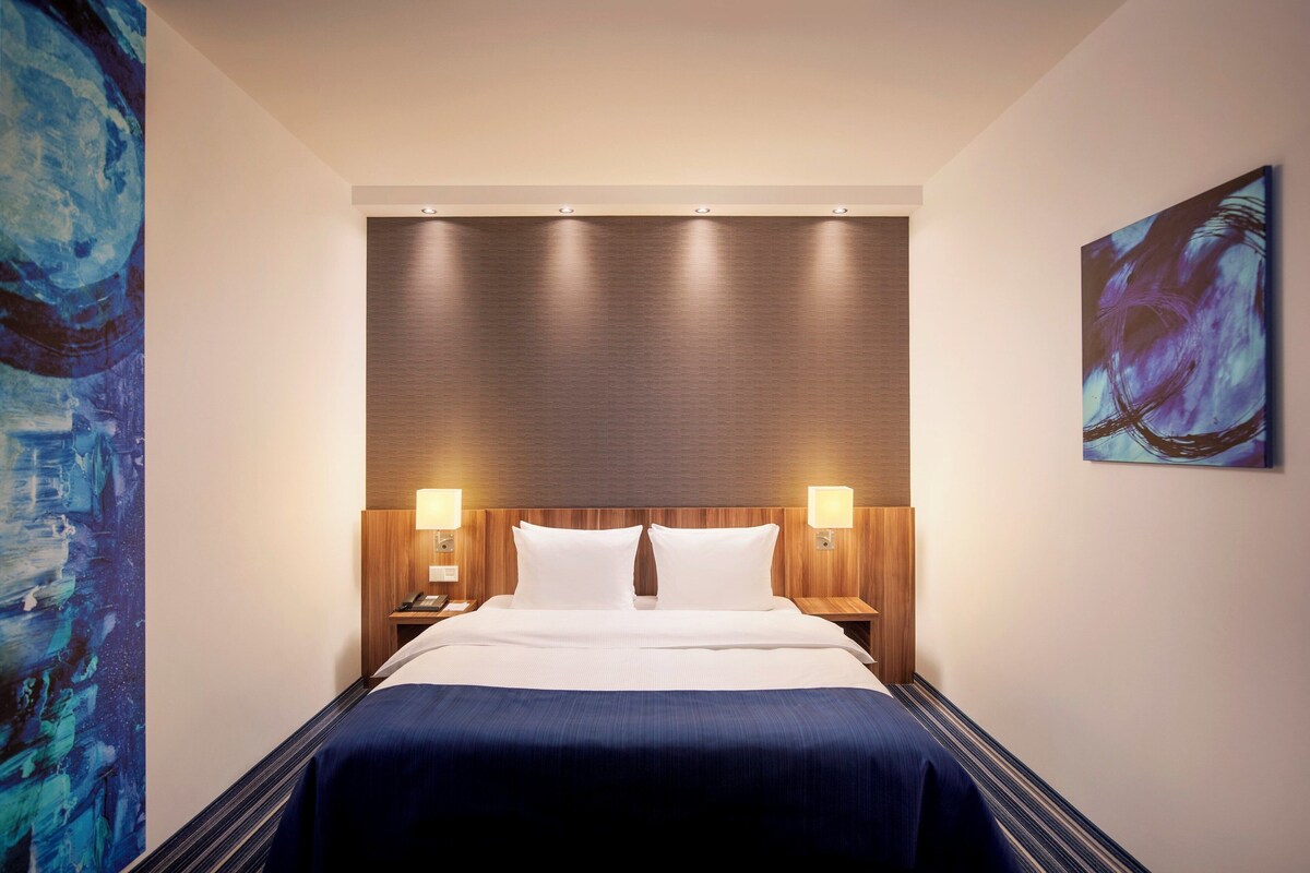 A hotel room with a large bed, two bedside lamps, a wooden headboard, and a large abstract painting on the wall.