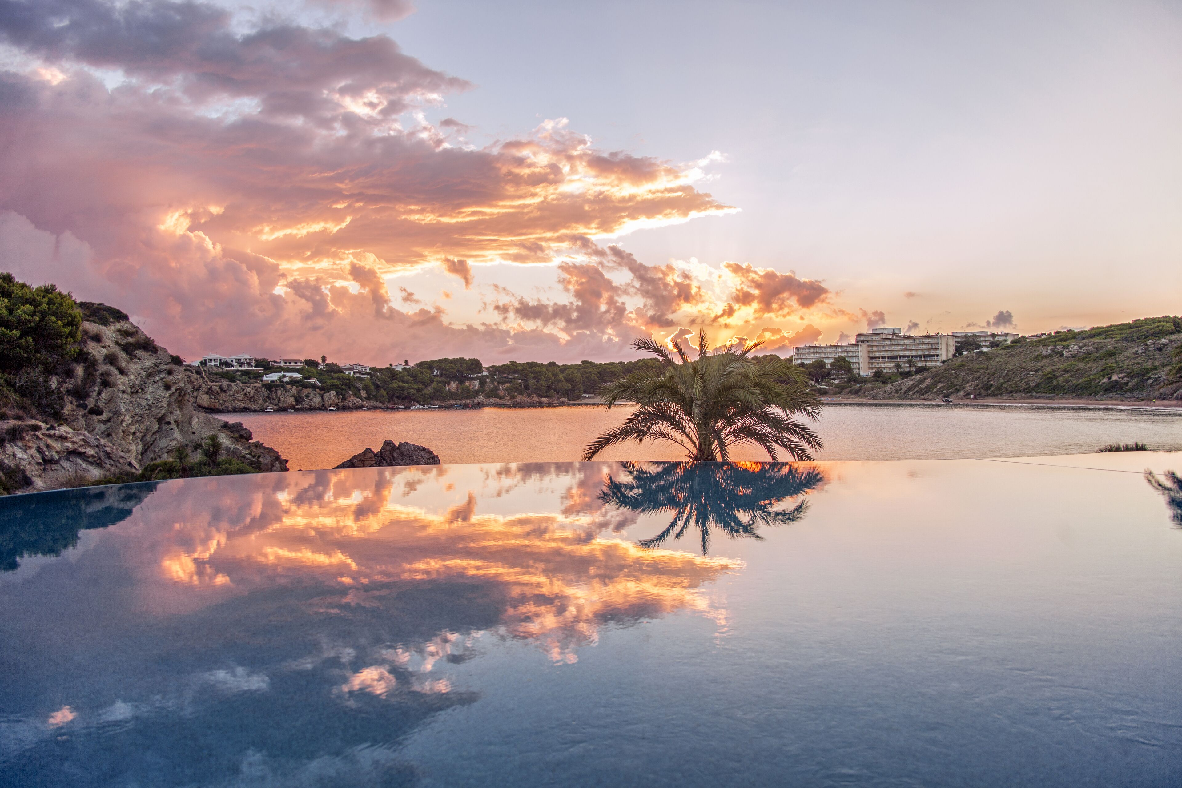 Vista despejada de una piscina con una palmera en el centro, rodeada de terreno rocoso y una masa de agua al fondo.