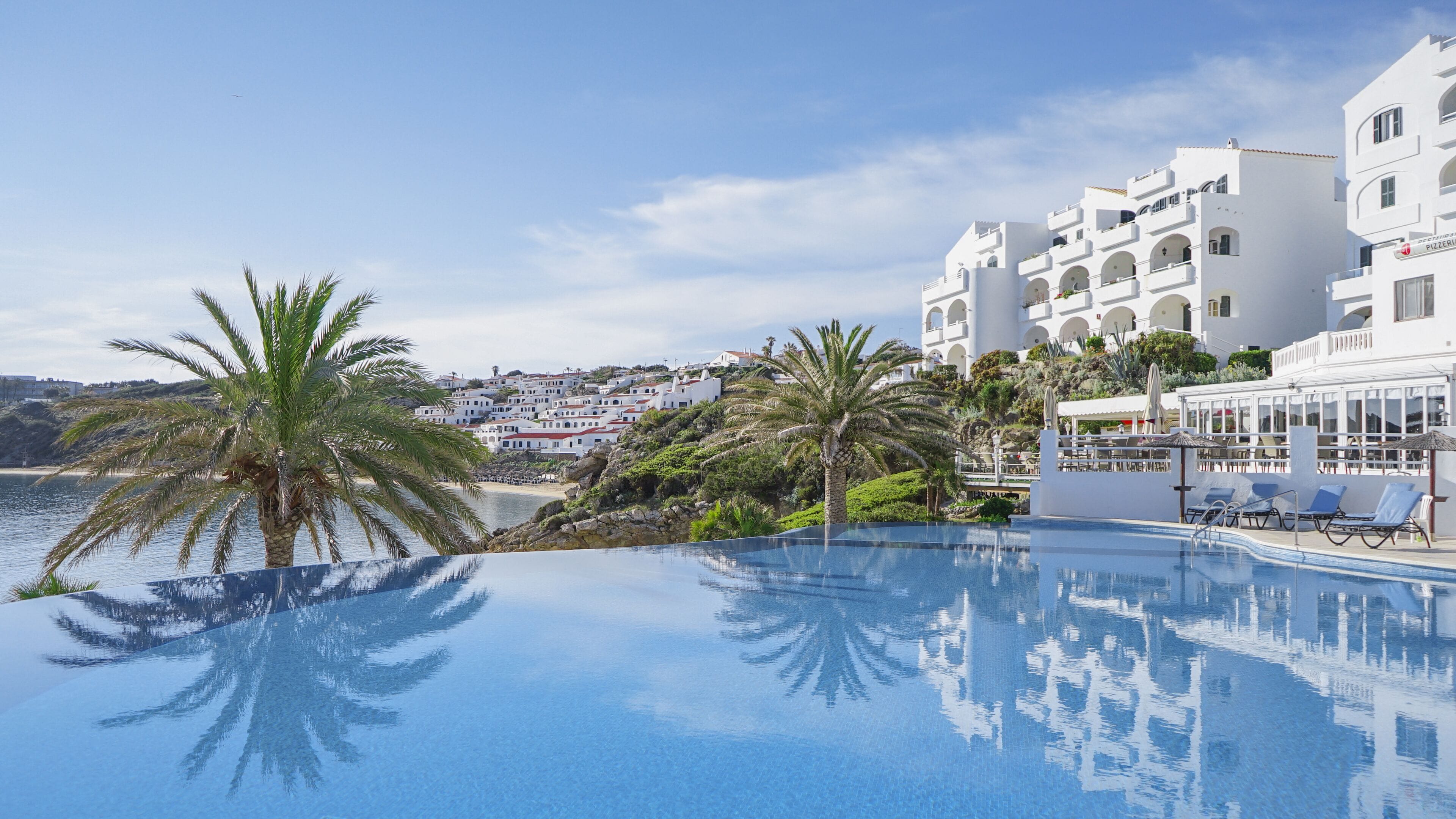 Una piscina infinita de color azul intenso con palmeras, una playa y edificios blancos con balcones.
