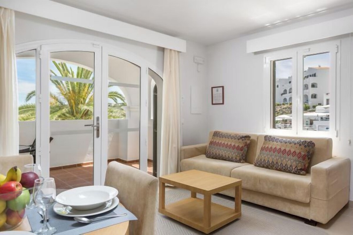 A living room with a sofa, a coffee table, and a dining table with plates and glasses. There is a sliding glass door leading to an outdoor area with a view of palm trees.