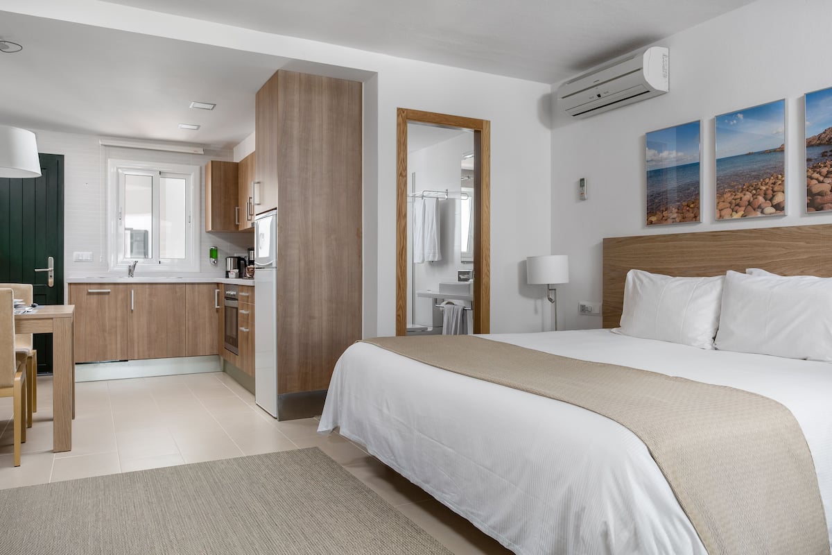 A modern hotel room with a large bed, a kitchen area, and a bathroom visible through an open door.