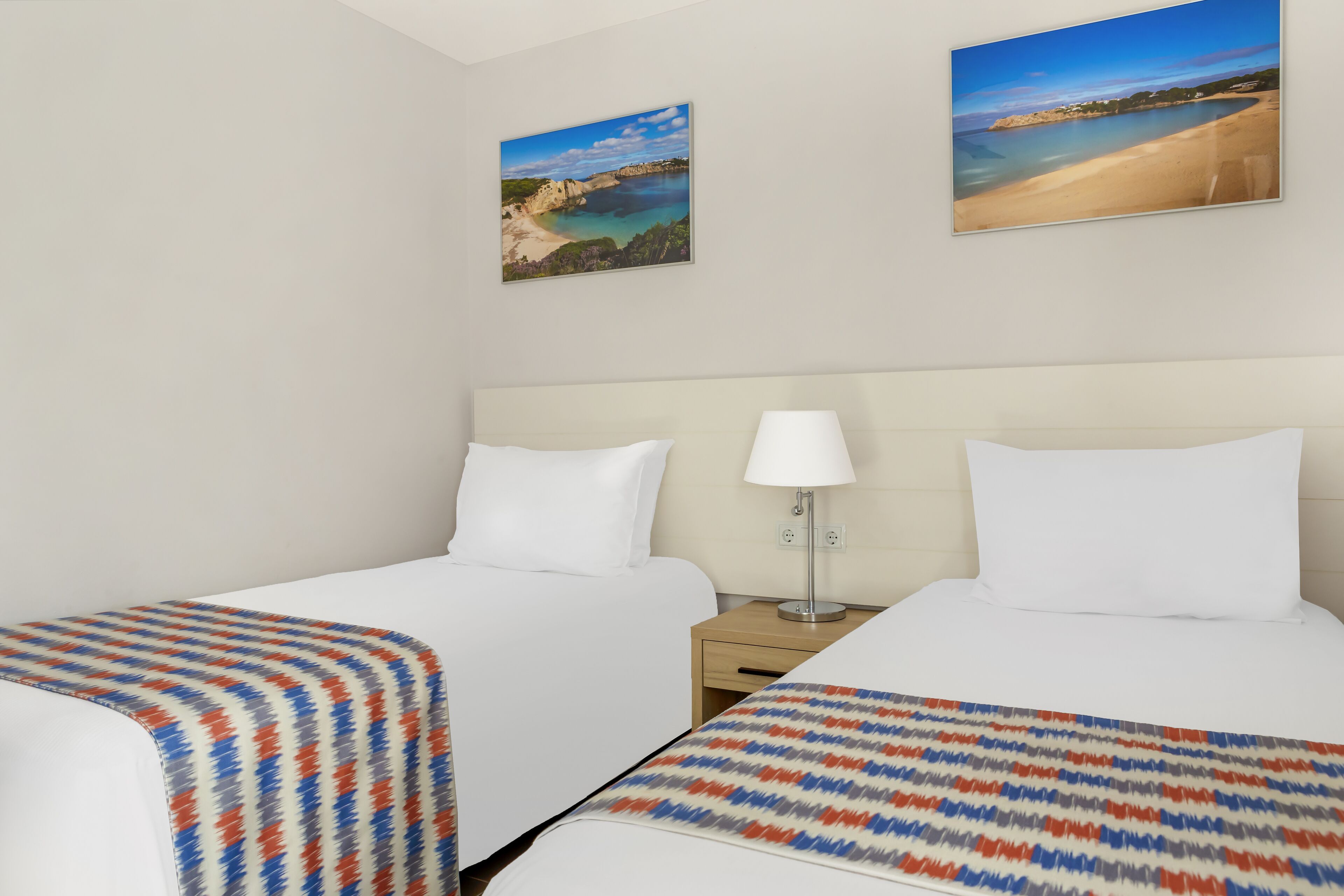A hotel room with two beds, a nightstand with a lamp, and two beach view paintings on the wall.