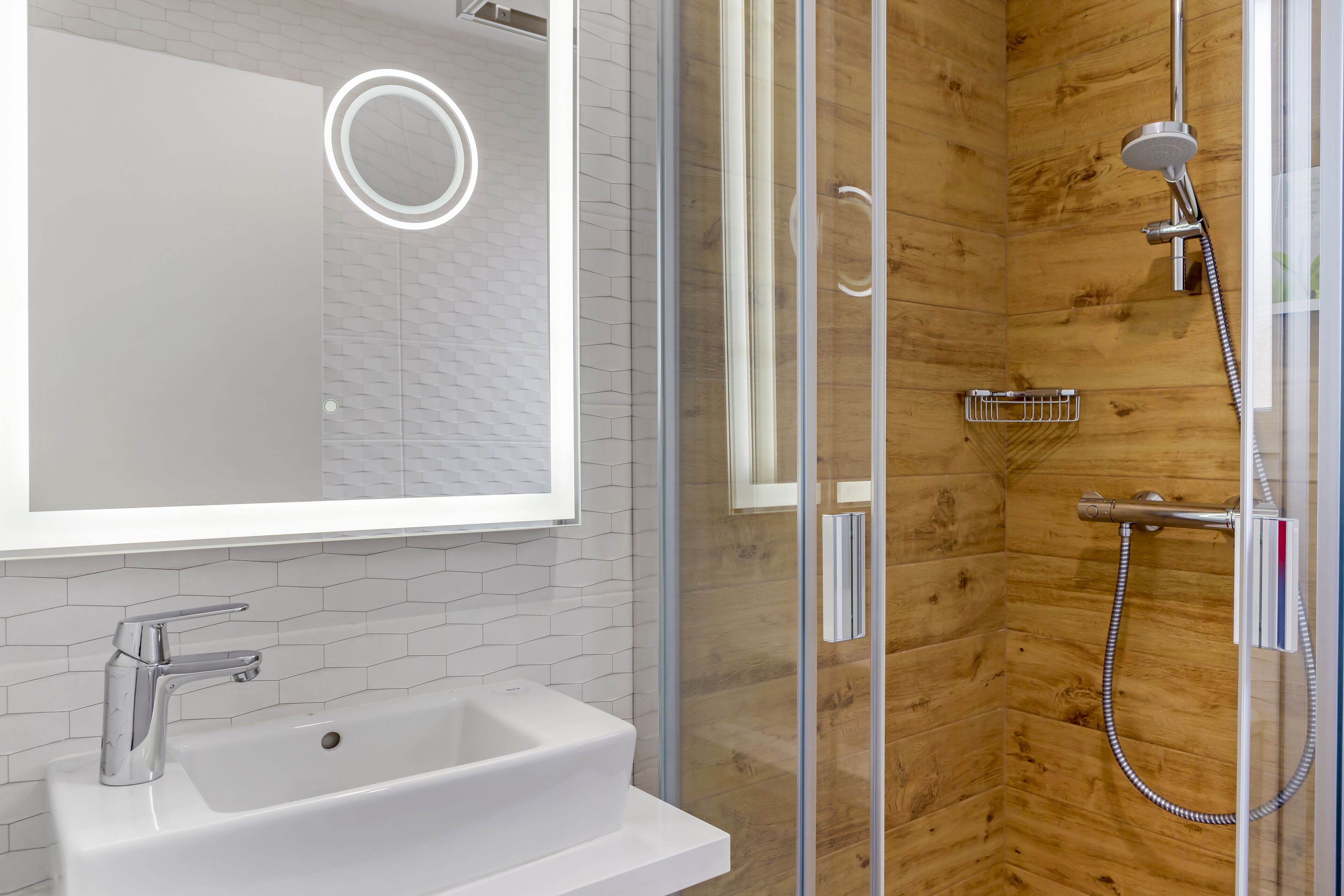 Une salle de bain moderne avec une douche à parois de verre, un lavabo blanc doté d’un robinet chromé et un miroir avec un éclairage circulaire.