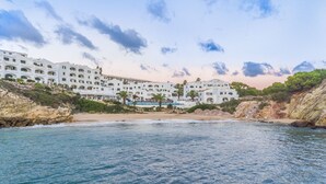 A coastal hotel with multiple white buildings, a sandy beach, and rocky cliffs. - White Sands Beach Club (Mercadal)