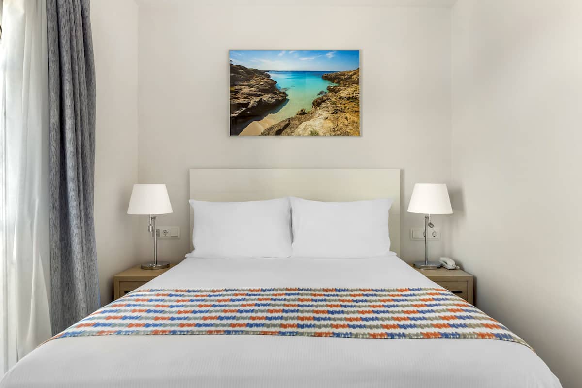 A hotel room with a bed, two bedside lamps, a painting of a coastal landscape, and a view of the sea.