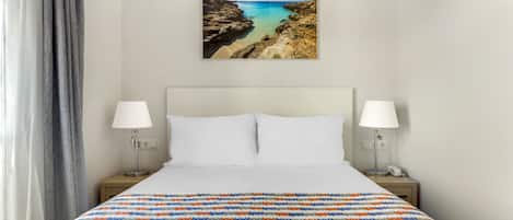 A hotel room with a bed, two bedside lamps, a painting of a coastal landscape, and a view of the sea.
