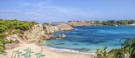 A coastal beach with clear blue waters, rocky cliffs, and green vegetation.