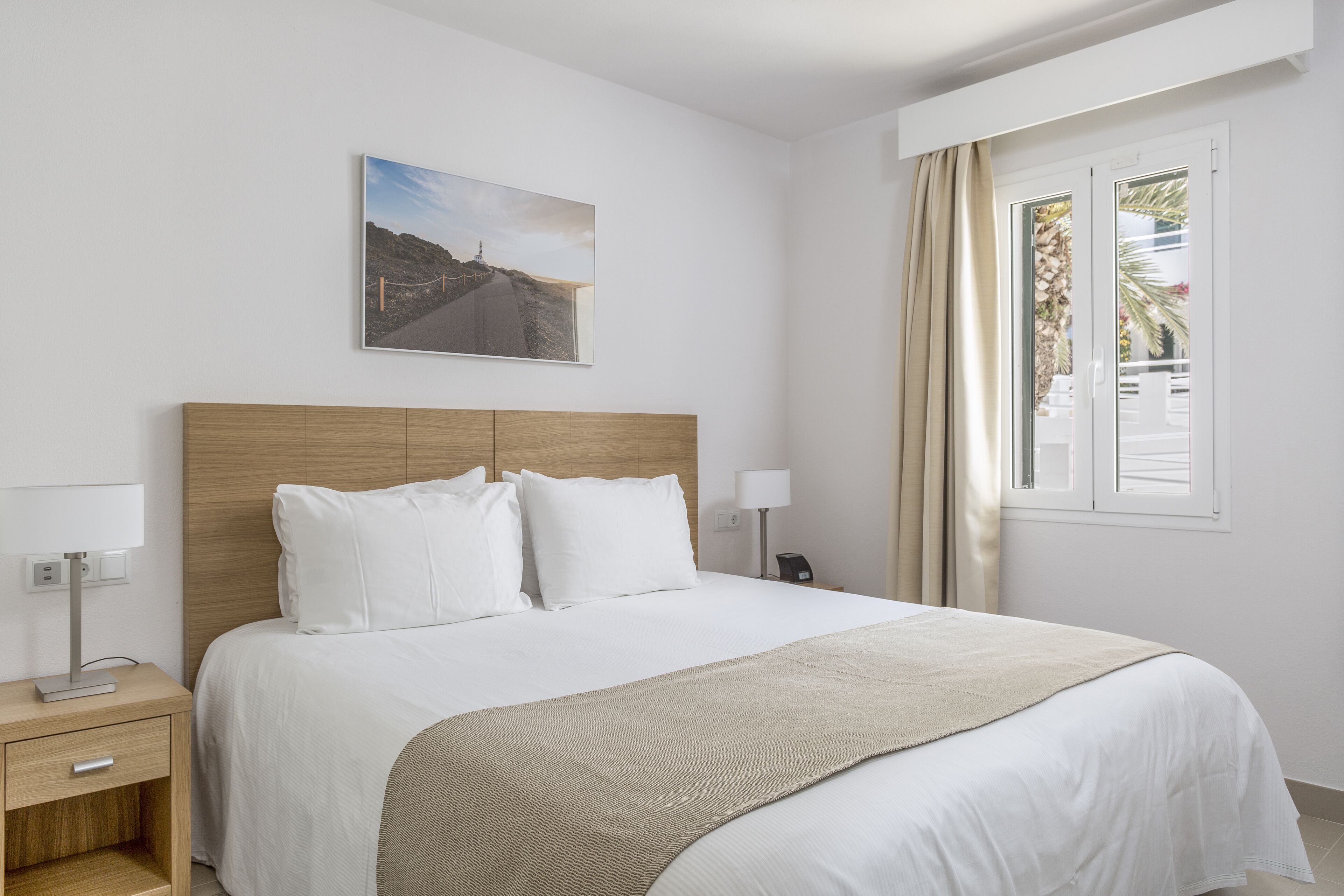 Una habitación de hotel con cabecera de madera, una cama grande con ropa blanca, un plaid color beige, una mesita de noche con lámpara y una ventana con cortinas.