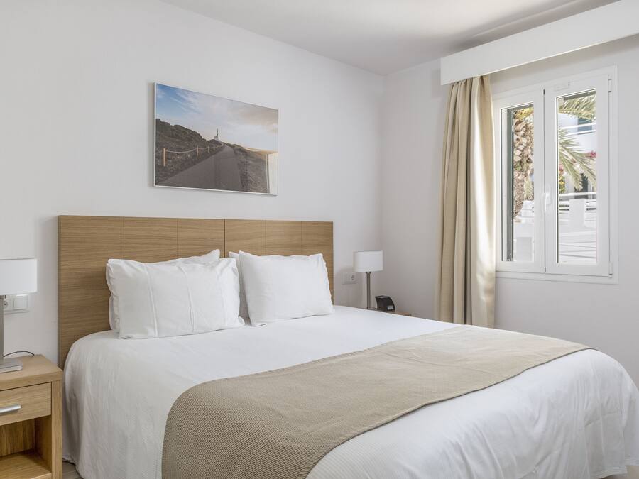 Una habitación de hotel con cabecera de madera, una cama grande con ropa blanca, un plaid color beige, una mesita de noche con lámpara y una ventana con cortinas.