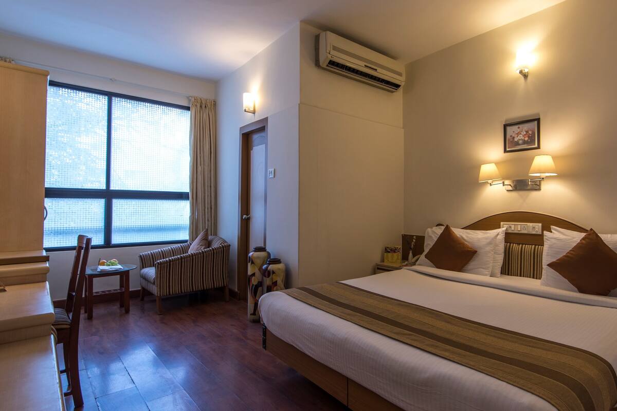 Habitación de hotel con cama, una silla, una mesita pequeña, aire acondicionado y ventana con cortinas.