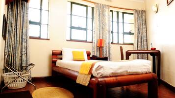 A room with a bed, a wooden bench, a small table, a lamp, a basket, and a window with curtains.