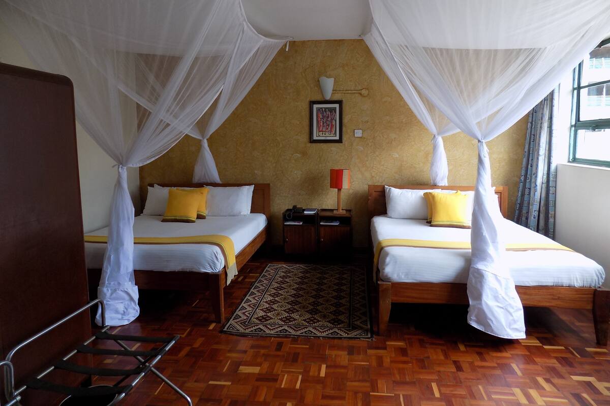 Ein Hotelzimmer mit zwei Betten, jeweils mit Baldachin, Holzboden, einem gemusterten Teppich und einem Nachttisch mit Lampe.