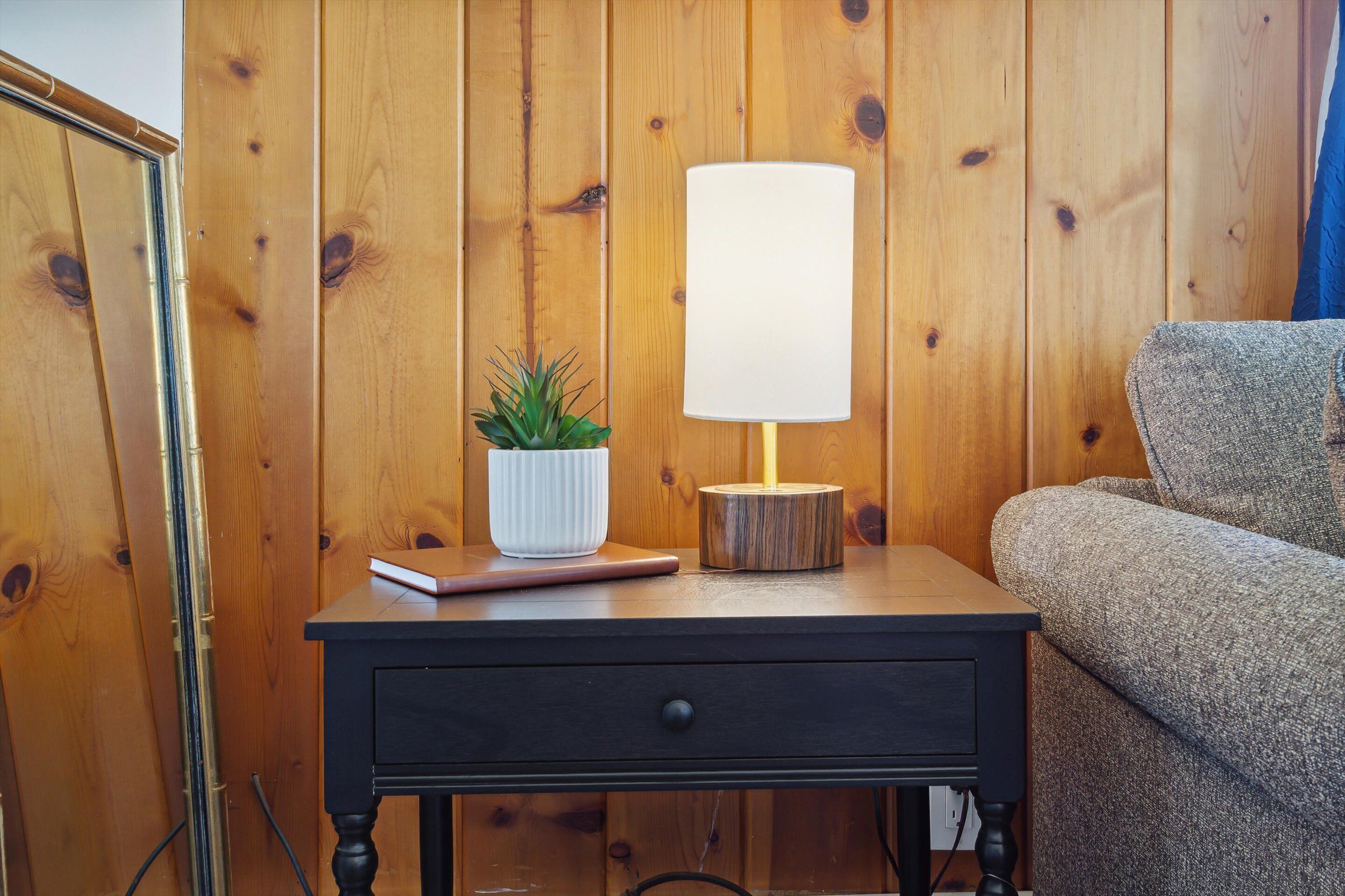 A wooden-paneled wall, a small wooden nightstand with a lamp and a plant, and a grey sofa.