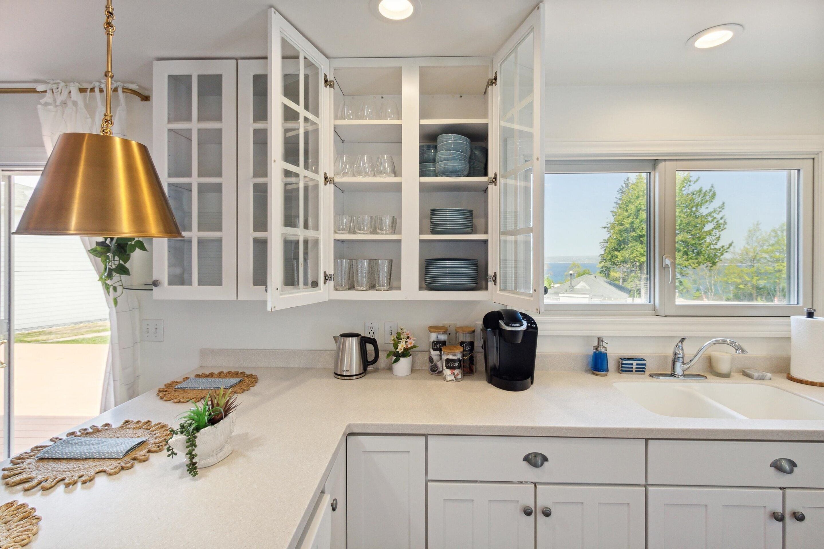 A kitchen with white cabinets, a coffee maker, a sink, and a view of the outdoors.