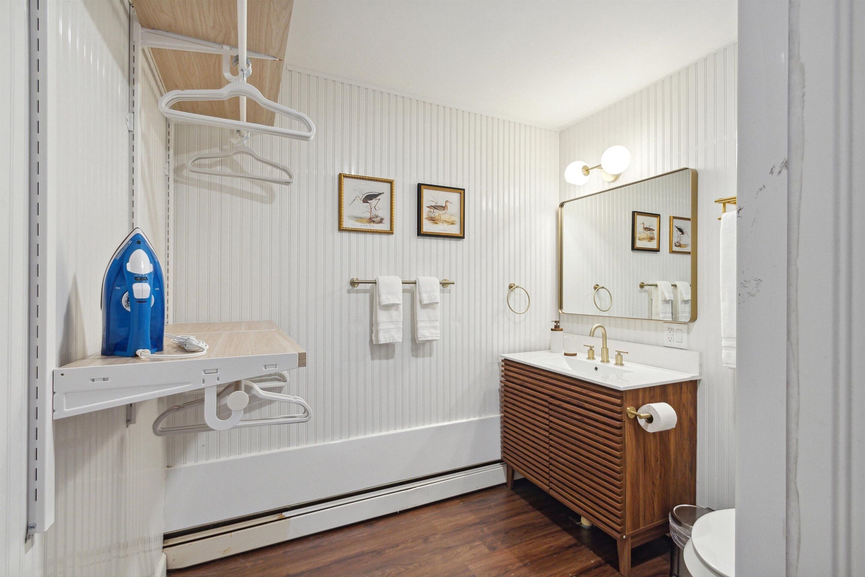 Une salle de bain avec un meuble-lavabo en bois, un lavabo blanc, un miroir avec éclairage intégré et deux tableaux encadrés accrochés au mur.