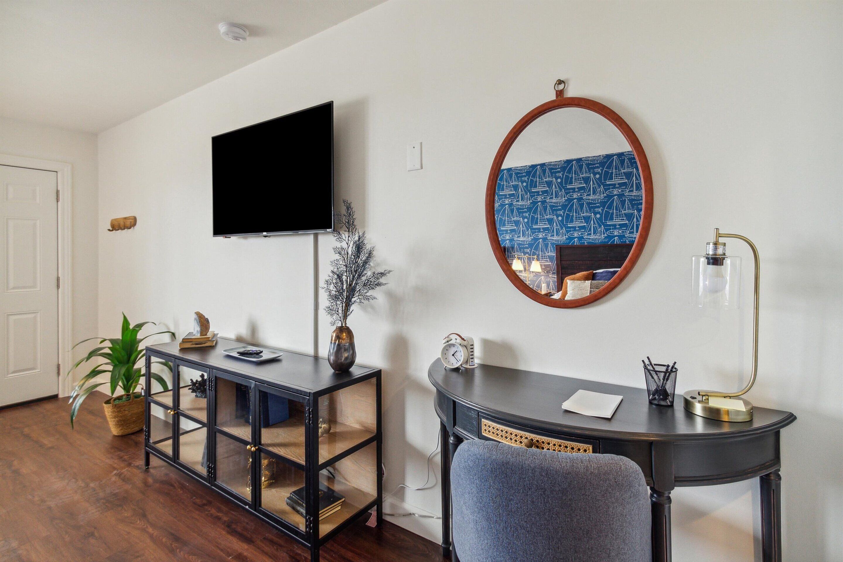 A room with a flat-screen TV mounted on the wall, a round mirror with a blue patterned frame, a dark wooden desk with a chair, and a potted plant.