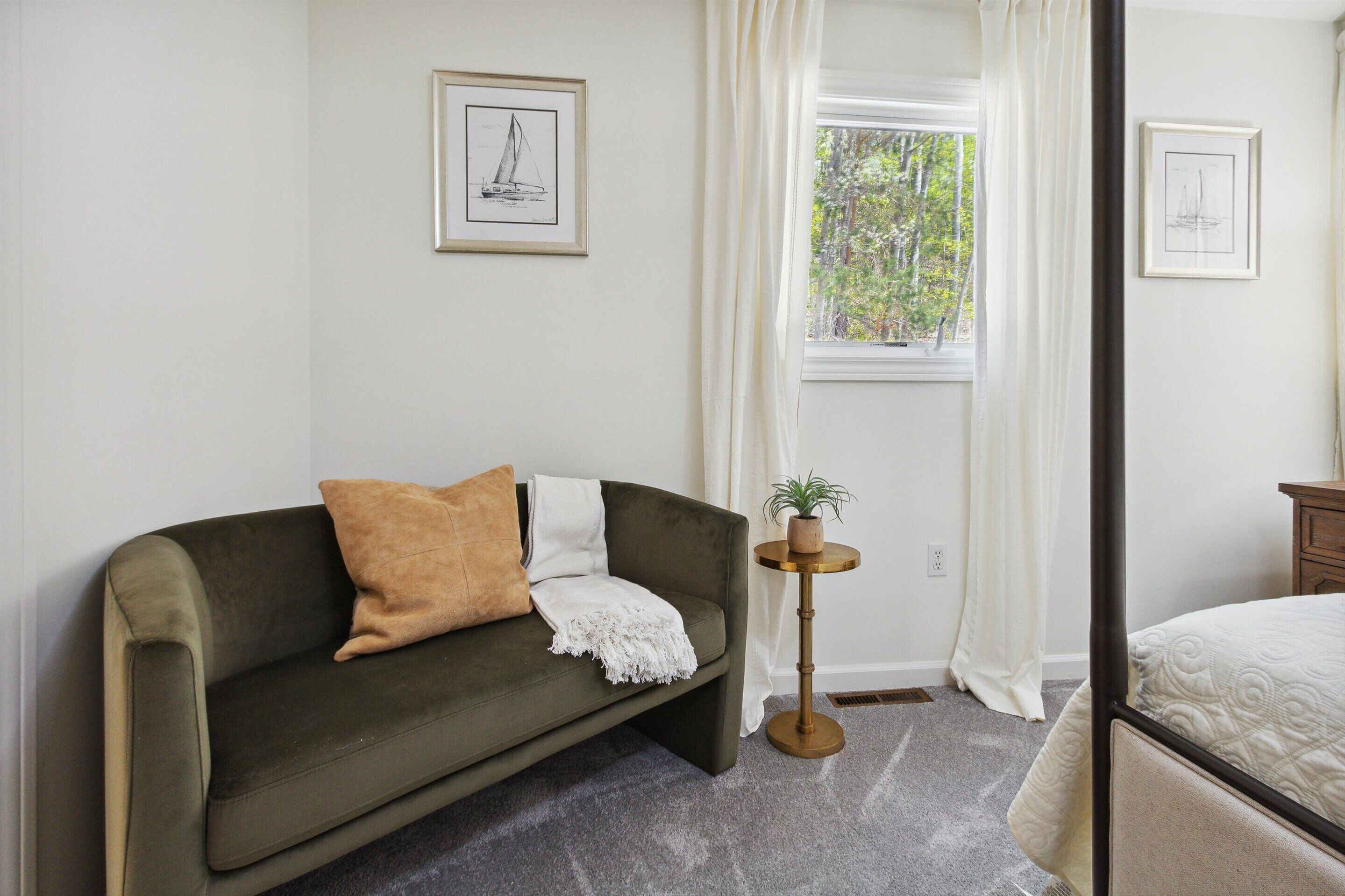 Quarto com sofá verde, uma mesa redonda pequena com uma planta, cama com colcha branca e janela com cortinas.