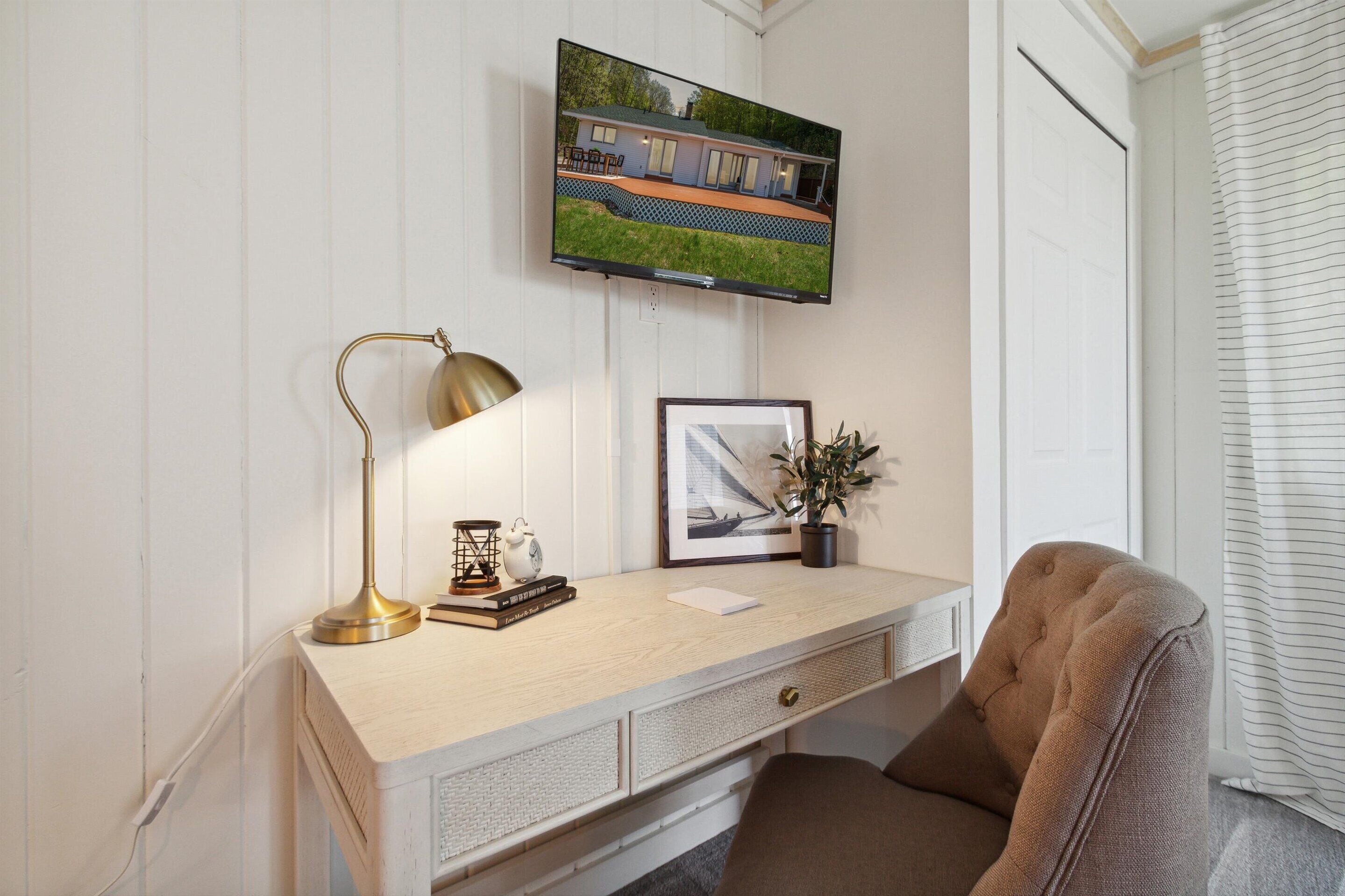 A neatly arranged workspace with a desk, chair, lamp, framed picture, and a television on the wall.