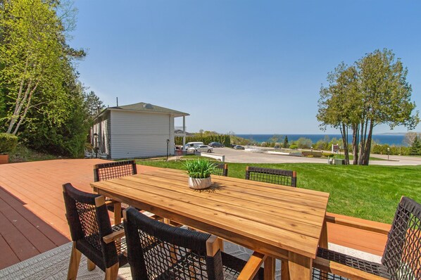 A wooden deck with a table and chairs, a potted plant, and a view of a house and the ocean.