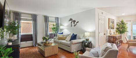 A spacious living room with a sofa, armchairs, and a coffee table. There are potted plants and a dining area visible.