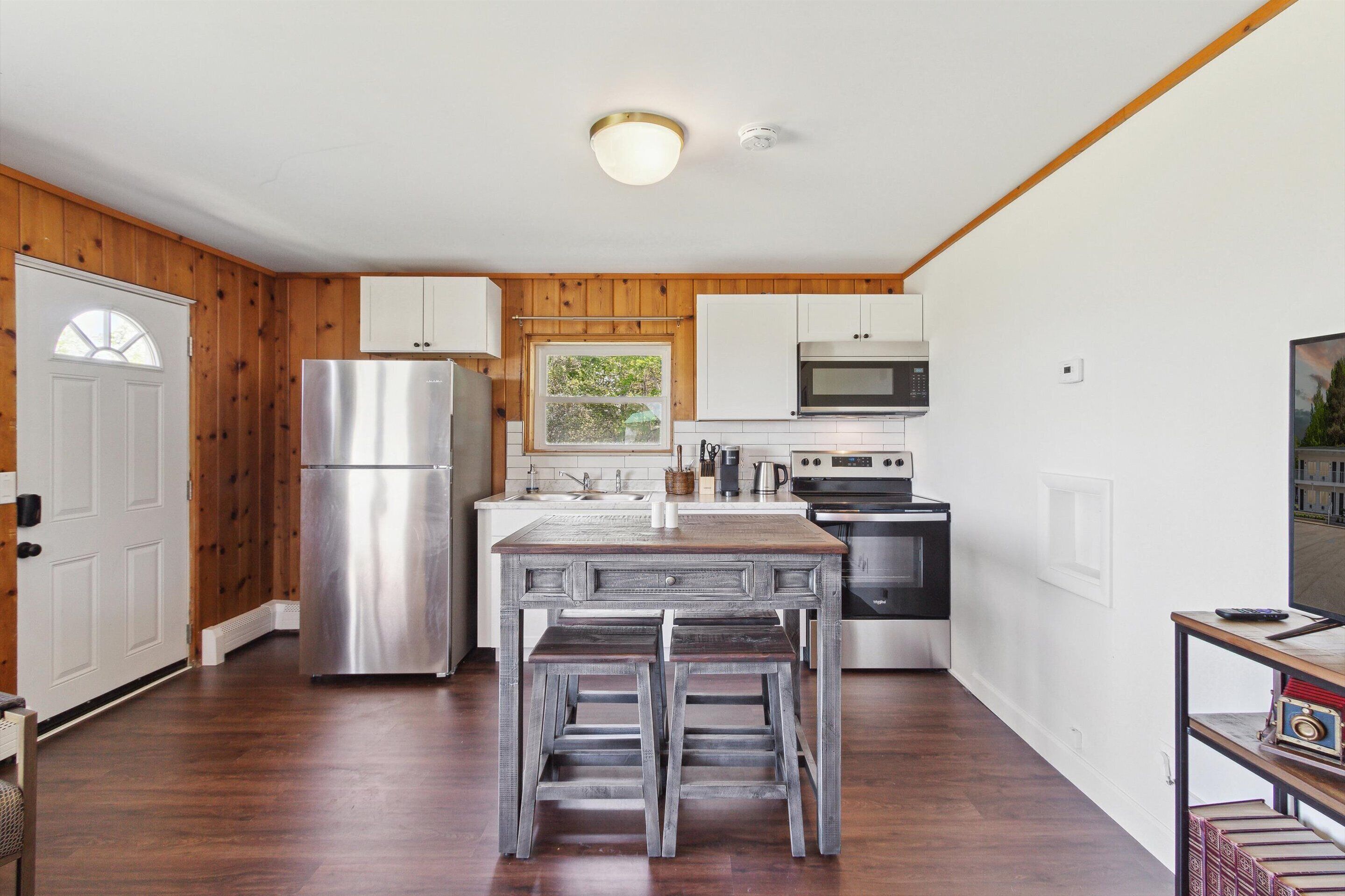 A kitchen with wooden walls, white cabinets, a stainless steel refrigerator, a microwave, and a stove. There is a window with a view of greenery.