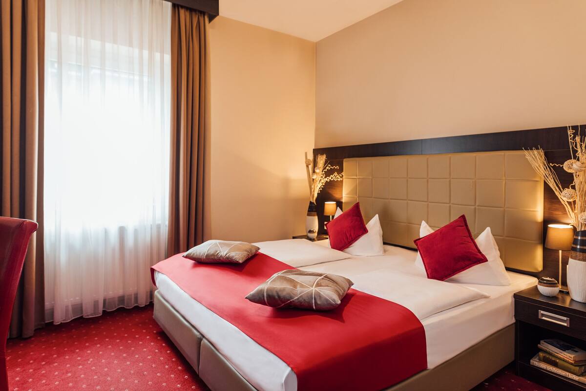 A hotel room with a large bed, two bedside tables, a nightstand, a red carpet, and a window with sheer curtains.