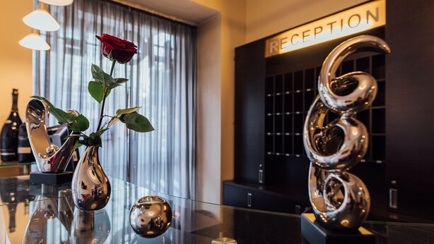 Ein Empfangsbereich mit einem Glastisch, auf dem eine Rose in einer Vase, eine dekorative Metallskulptur und ein Schild mit der Aufschrift 'RECEPTION' stehen.