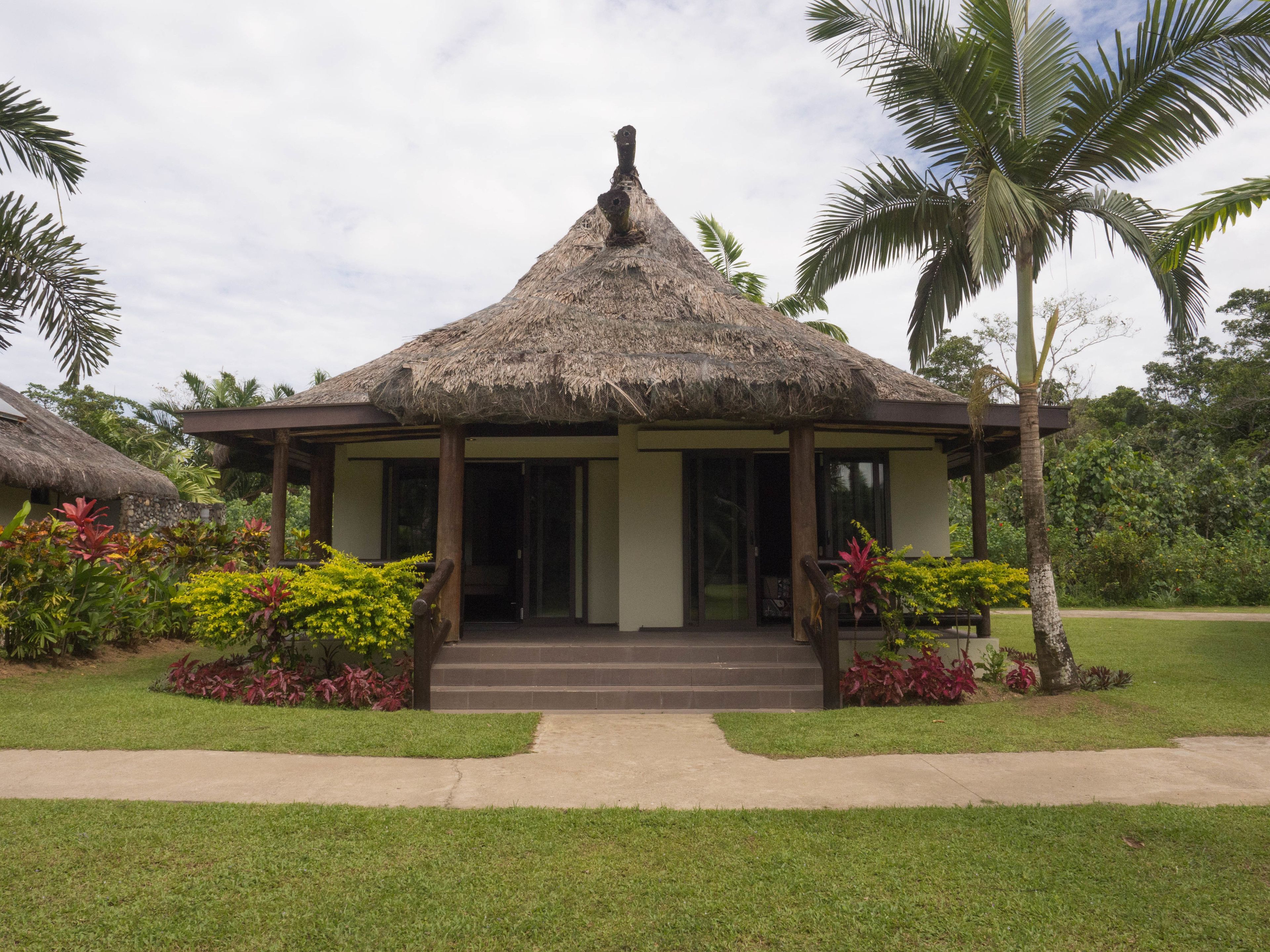 A thatched-roof structure with a covered entrance and steps leading up to it, surrounded by lush greenery and palm trees.