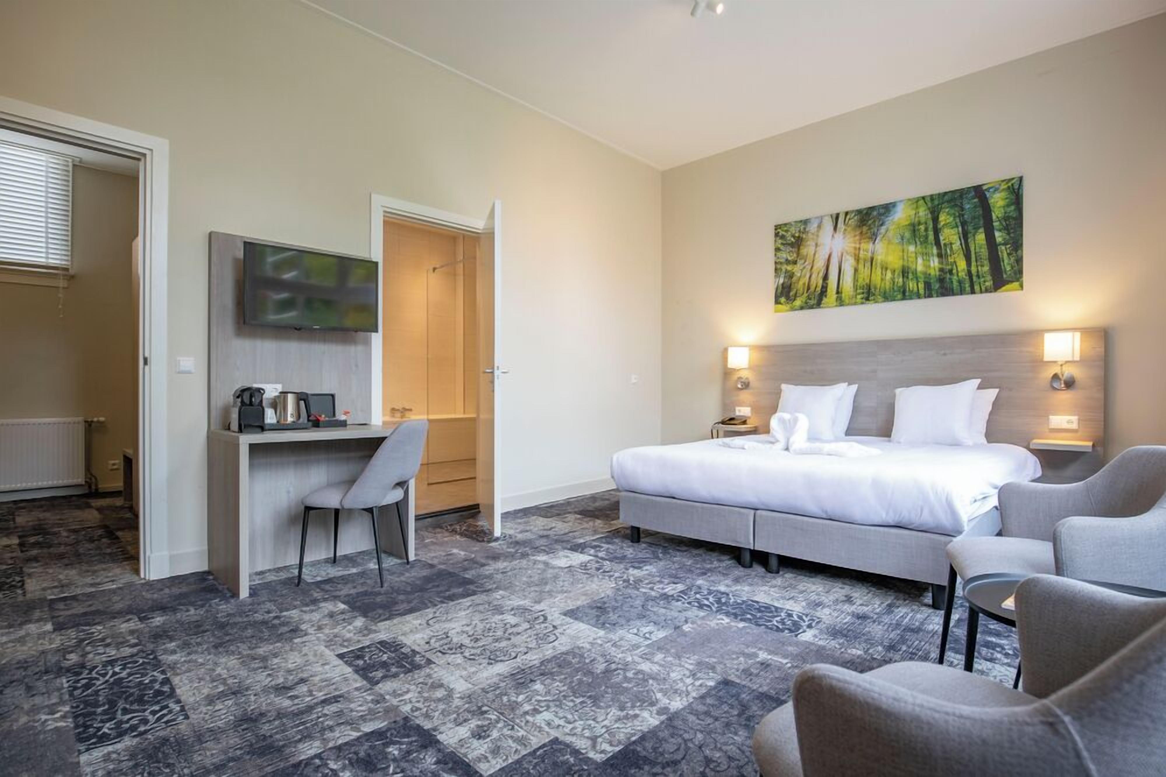 A hotel room with a bed, a desk, a chair, a painting on the wall, and a patterned carpet.