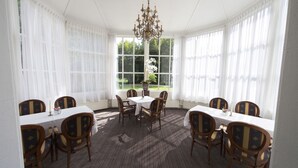 Een kamer met een ronde tafel en stoelen, grote ramen met witte gordijnen en een kroonluchter.