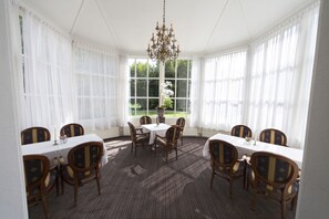 Une pièce avec une table ronde et des chaises, de grandes fenêtres avec des rideaux blancs et un lustre.