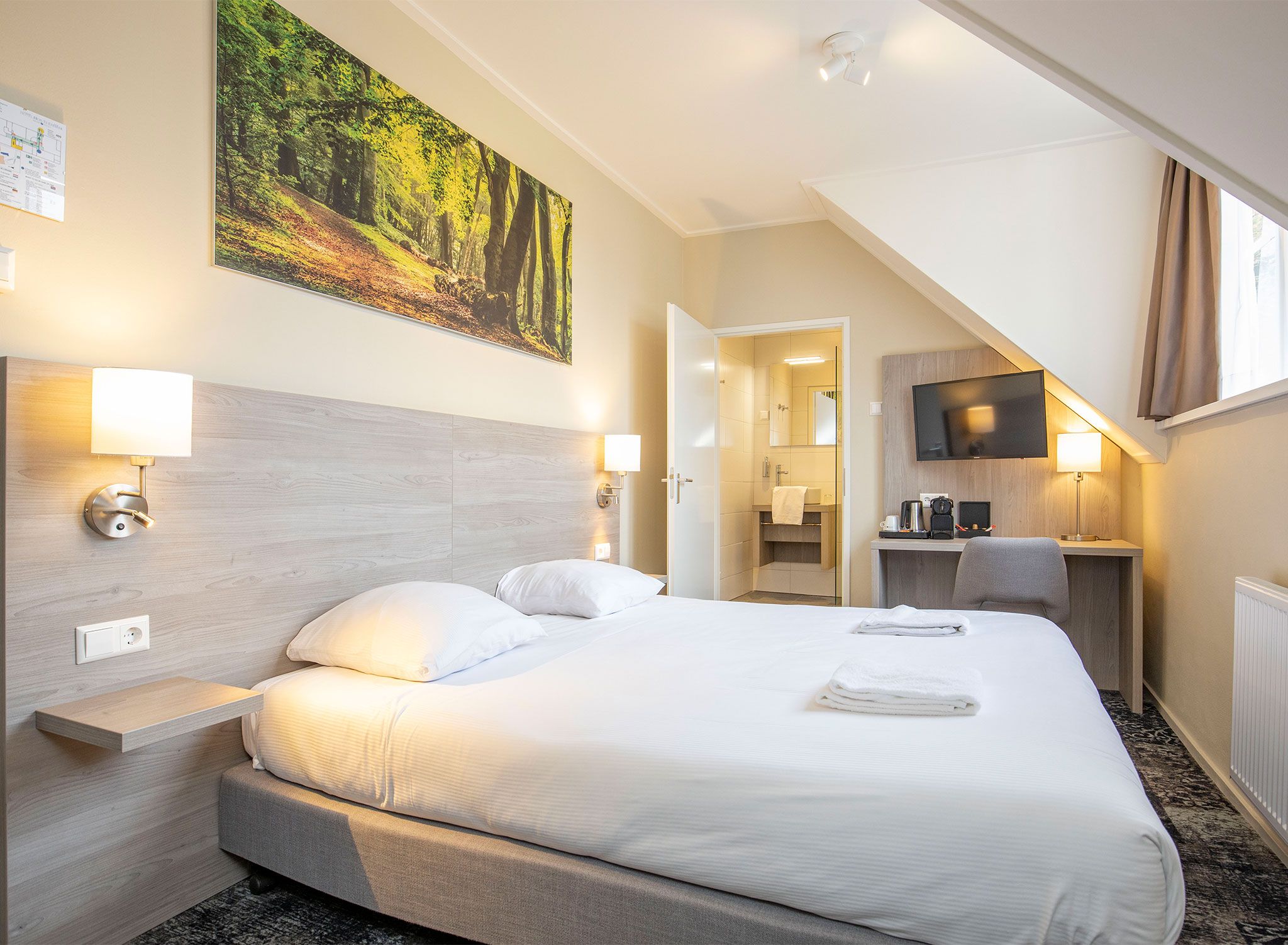 A hotel room with a bed, bedside lamps, a flat-screen TV, and a painting of a forest.
