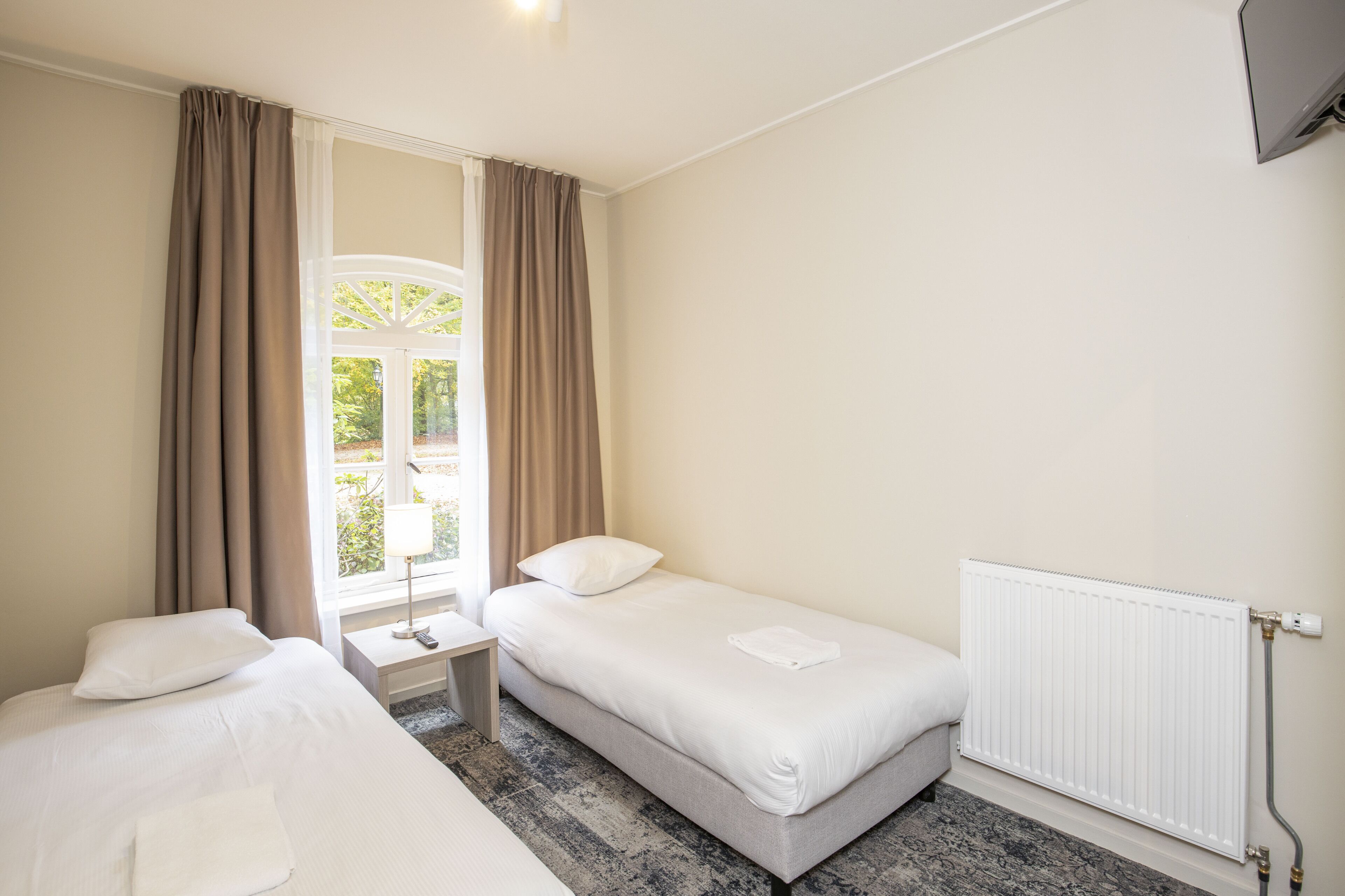 A hotel room with a bed, a small bedside table, a radiator, and a window with curtains.