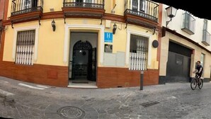 A narrow street with buildings on both sides. There is a building with a yellow facade and a black gate in the center. - Hostal Paco's (Seville)