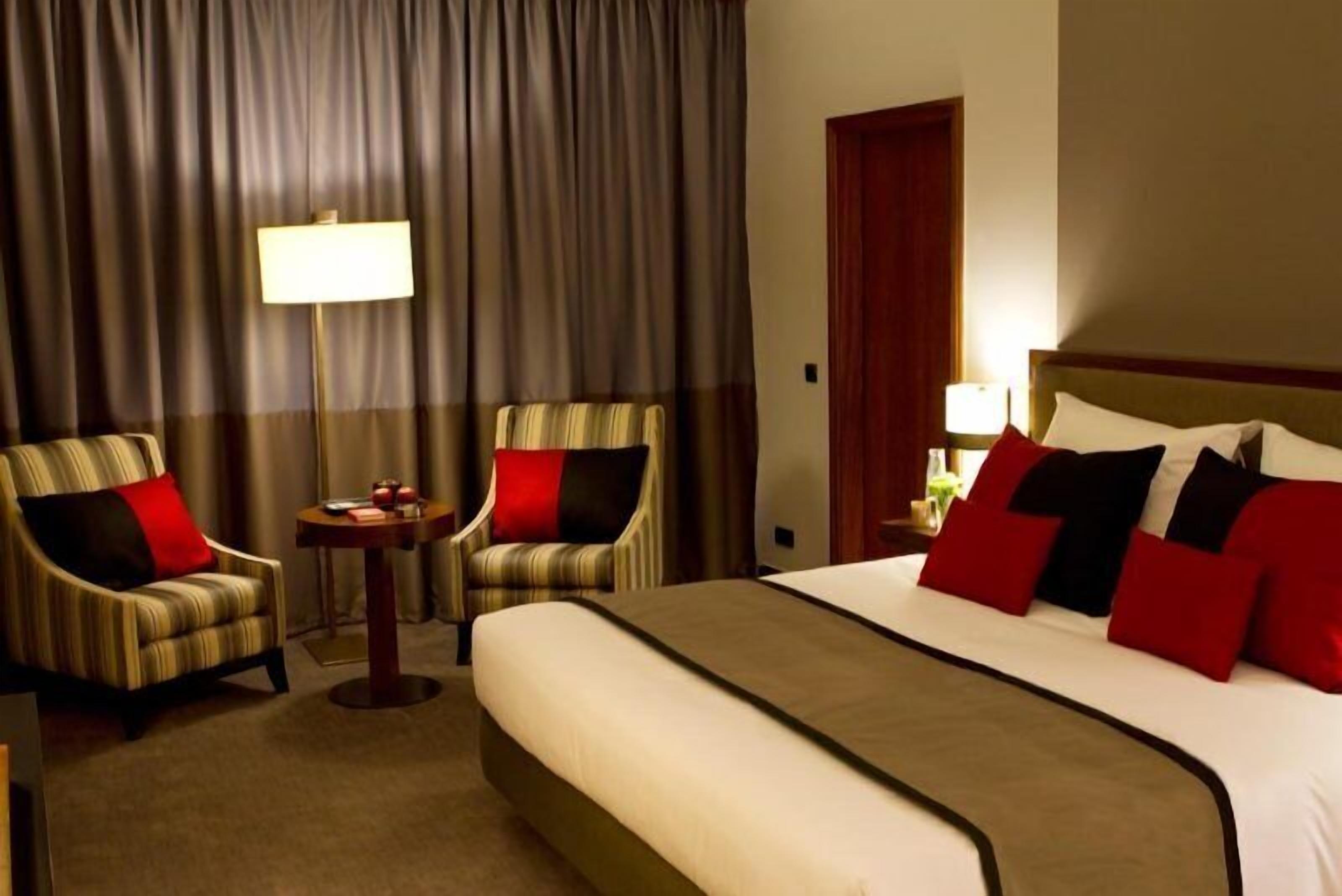 Une chambre d’hôtel avec un grand lit, deux fauteuils, une petite table, une lampe et une porte.
