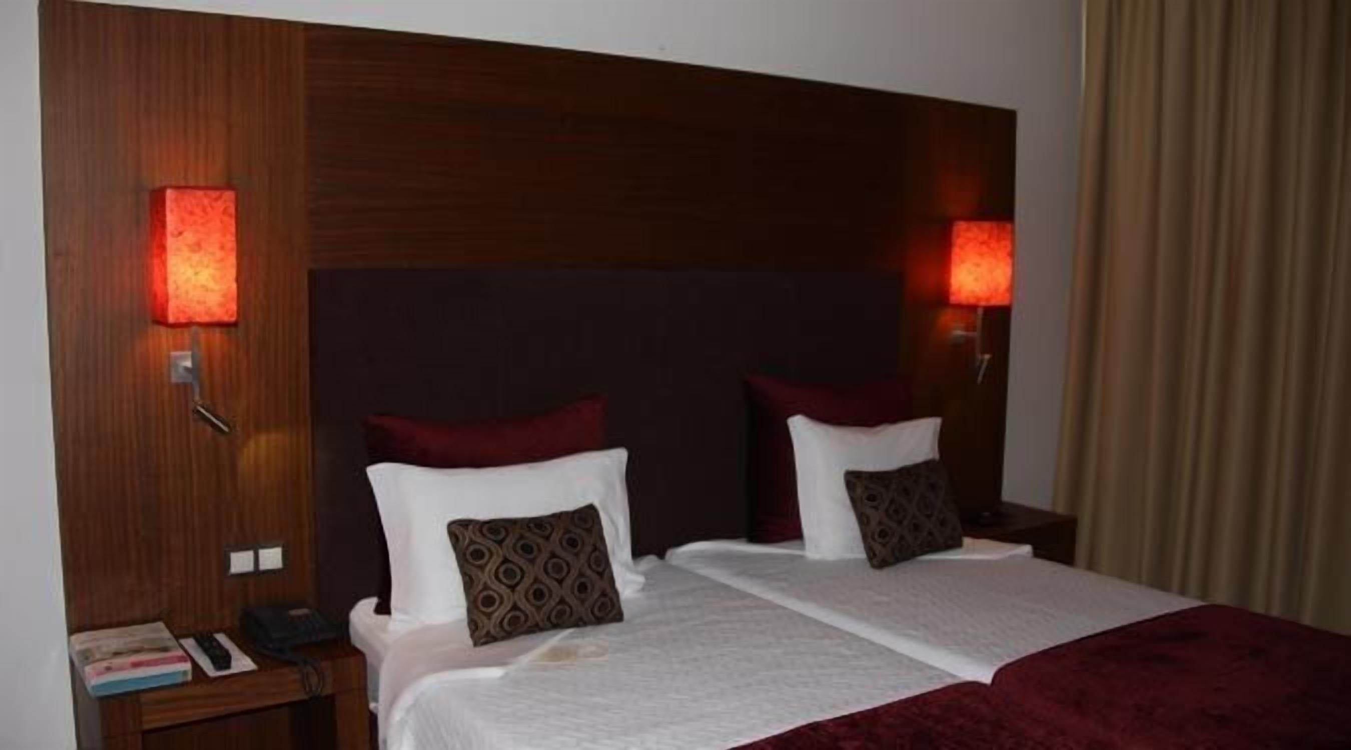 A hotel room with a wooden headboard, two beds with white linens and brown pillows, a nightstand with a phone and books, and two wall-mounted lamps.
