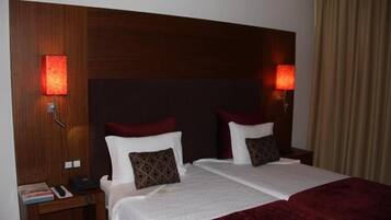 A hotel room with a wooden headboard, two beds with white linens and brown pillows, a nightstand with a phone and books, and two wall-mounted lamps.