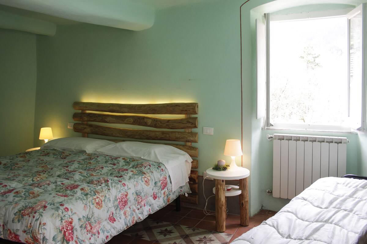 Un dormitorio con cabecera de madera, colcha estampada, mesita de noche con lámpara y ventana con vistas a árboles.