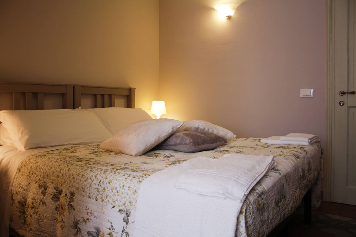 Una cama bien hecha con una colcha estampada y almohadones, una lámpara de noche y una puerta a la derecha.
