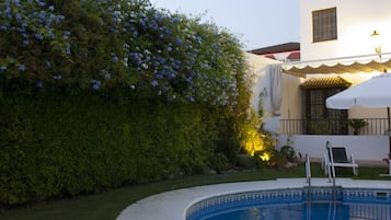 Une piscine avec un parasol blanc, un bâtiment blanc doté d’un toit en tuiles et une haie verdoyante ornée de fleurs bleues.