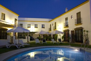 Un área de piscina con sillones de descanso y sombrillas, rodeada por un edificio con balcones y ventanas.