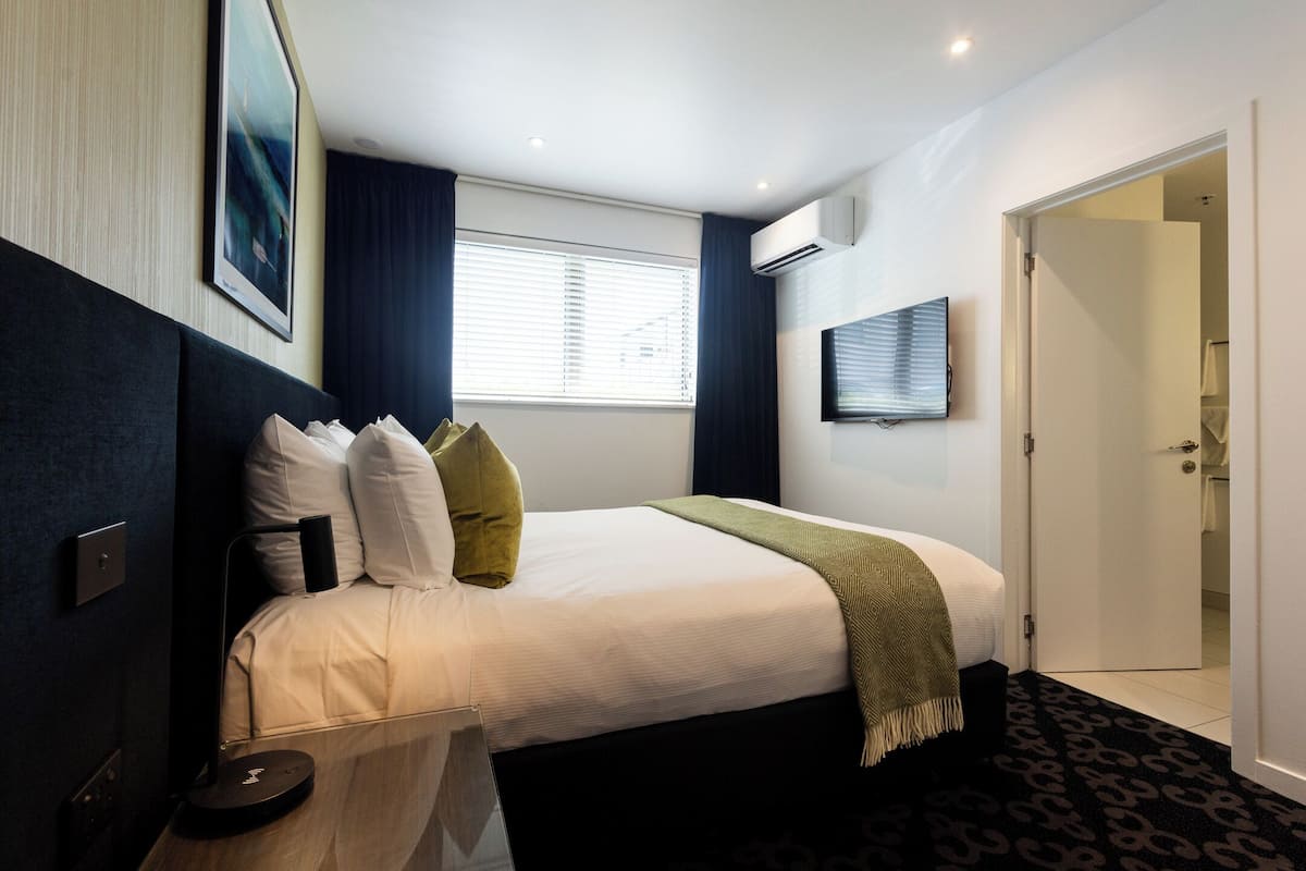 A modern hotel room with a neatly made bed, a flat-screen TV mounted on the wall, and a door leading to a bathroom.